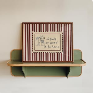 Puede incluir: Una impresión enmarcada con un borde marrón y un fondo a rayas granate y blanco. La impresión presenta un rectángulo beige con una ilustración de una casa y el texto "it feels so good to be home". La impresión se muestra en una estantería verde y beige.