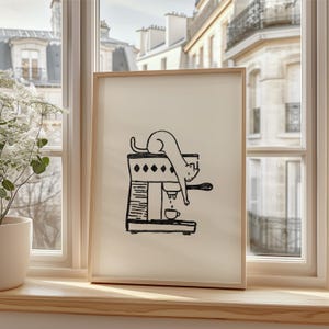 Puede incluir: Una impresión artística enmarcada con una ilustración en blanco y negro de un gato en una máquina de café. El gato está tumbado sobre la máquina, con una pequeña taza debajo. La impresión se muestra en un alféizar de madera.