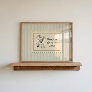 Op de afbeelding: Een ingelijste print met een lichtbeige achtergrond en een houten frame. De print heeft een postzegelontwerp met een huis, bergen en de tekst "there's no place like home". Het frame staat op een houten plank.