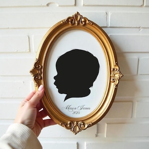 May include: Oval gold-colored picture frame with a black silhouette of a child's profile on a white background. The frame has ornate detailing. The name "Mason James" and the year "2025" are written in script below the silhouette.