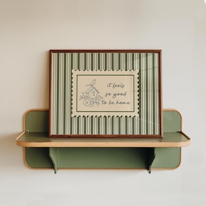 Puede incluir: Un cuadro enmarcado con una ilustración de una casa para pájaros y el texto "it feels so good to be home". El cuadro tiene un marco marrón y está sobre un fondo a rayas verdes y beige. Descansa sobre una estantería verde y beige.