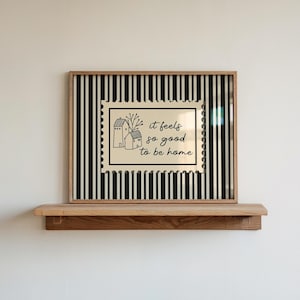 Peut inclure: Une impression d'art encadrée avec un fond à rayures noires et blanches. L'impression présente un motif beige en forme de timbre avec deux illustrations de maisons et le texte "it feels so good to be home". Le cadre est en bois naturel, et l'art est exposé sur une étagère en bois.