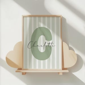 May include: A framed print featuring a large green letter "C" and the name "Charlotte" in elegant script. The background has vertical stripes in light green and white. The print is displayed on a wooden shelf with cloud-shaped accents.