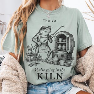 May include: A sage green t-shirt featuring a black ink-style illustration of a frog wearing an apron, holding a tool, and standing next to a kiln. The text reads: That's it. You're going in the KILN. The shirt has a crew neck and short sleeves.