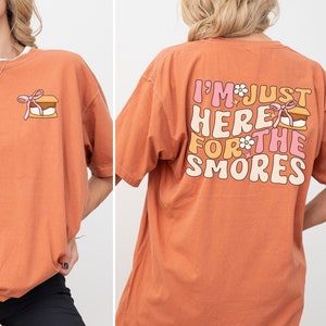 Op de afbeelding: Een oranje T-shirt met een afbeelding op de voorkant van een s'more en de tekst "I'm just here for the s'mores" op de achterkant.