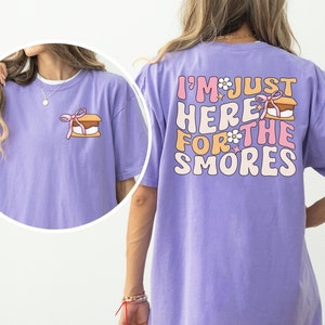 Op de afbeelding: Een paars T-shirt met de tekst "I'm just here for the s'mores" in roze en witte letters met een s'mores-afbeelding. Het shirt heeft een ontspannen pasvorm.