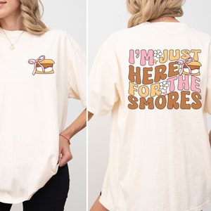 Op de afbeelding: Een cr&egrave;mekleurig T-shirt met een afbeelding van een s'more op de voorkant en de tekst "I'm just here for the smores" op de achterkant.