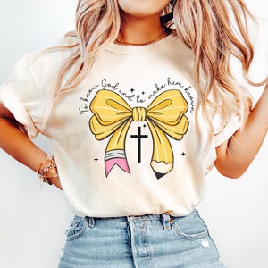 May include: A white t-shirt with a yellow pencil bow and a cross. The text on the shirt reads "To know God and to make him known."