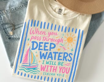 Könnte beinhalten: Ein cremefarbenes T-Shirt mit einem Grafikdruck, der ein Segelboot, eine Sonne und den Text "When you pass through DEEP WATERS I WILL BE WITH YOU ISAIAH 43:2" in Blau und Pink zeigt, kombiniert mit einer hellblauen Jeans und einem beigen Hut.