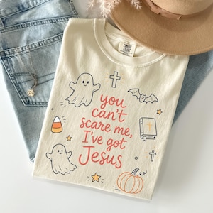 May include: A cream-colored t-shirt featuring the red text 'You can't scare me, I've got Jesus' surrounded by illustrations of two ghosts, a cross, a bat, a Bible, a pumpkin, candy corn, and stars, paired with light blue denim jeans and a tan hat.