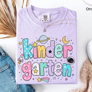 May include: An orchid-colored t-shirt featuring the word Kindergarten in large, multi-colored, patterned bubble letters. The design includes space-themed graphics such as a planet with rings, a rocket ship, an astronaut helmet, and stars.
