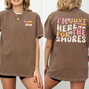 Op de afbeelding: Een bruin t-shirt met een afbeelding van een s'more op de voorkant en de tekst "I'm just here for the smores" op de achterkant.
