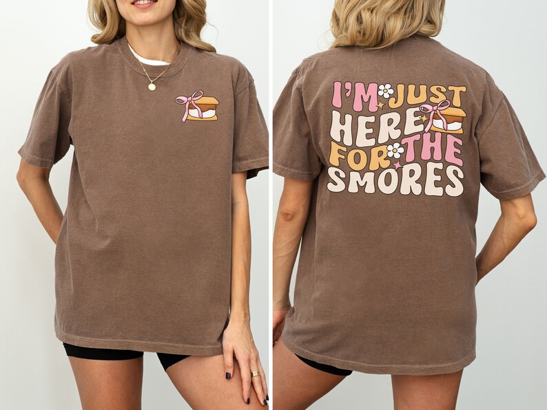 Op de afbeelding: Een bruin t-shirt met een afbeelding van een s'more op de voorkant en de tekst "I'm just here for the smores" op de achterkant.