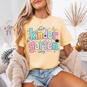May include: A pale yellow short-sleeve t-shirt featuring the word Kindergarten in large, colorful, patterned bubble letters. The design includes space-themed graphics such as a planet, a rocket, a crescent moon, stars, and an astronaut helmet.