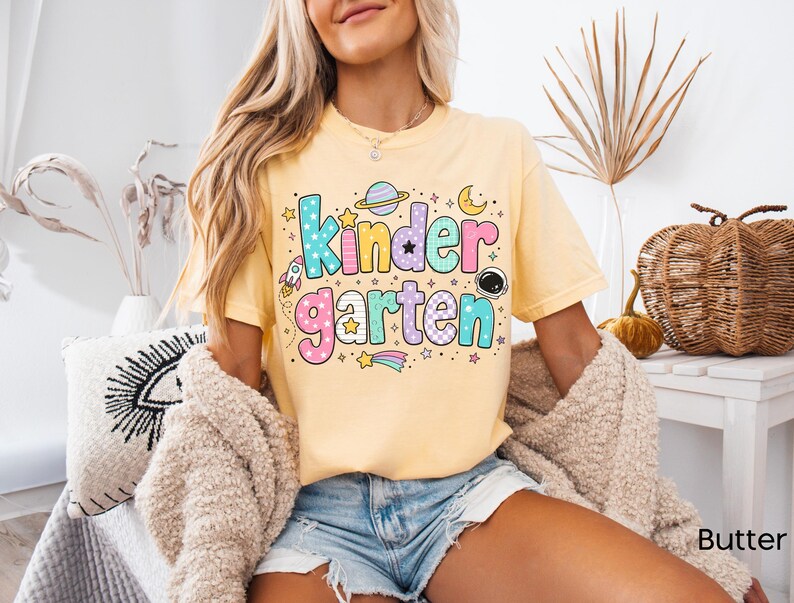 May include: A pale yellow short-sleeve t-shirt featuring the word Kindergarten in large, colorful, patterned bubble letters. The design includes space-themed graphics such as a planet, a rocket, a crescent moon, stars, and an astronaut helmet.
