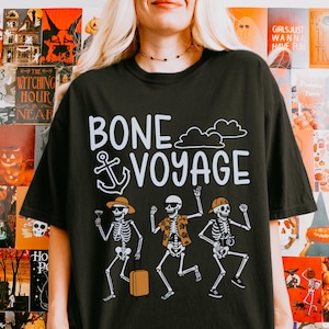May include: Black t-shirt with the phrase "BONE VOYAGE" and a graphic of dancing skeletons. One skeleton holds a cocktail, another a suitcase, and the third a camera. The shirt has a relaxed fit.
