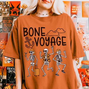 May include: An orange t-shirt with the words "BONE VOYAGE" and an anchor graphic. Below the text are three skeleton figures, one holding a cocktail, one with a suitcase, and one with a camera. The shirt has a crew neck and short sleeves.