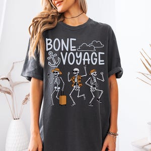 May include: Dark gray t-shirt with the phrase "BONE VOYAGE" and an anchor graphic. The shirt features a graphic of three skeleton figures, each with vacation-themed accessories, including sunglasses, hats, and luggage.