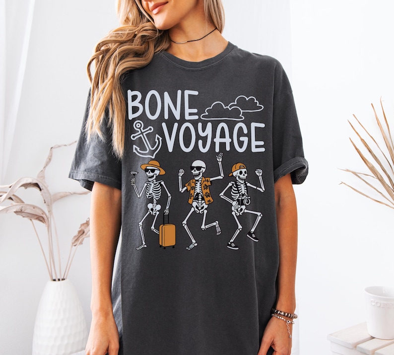 May include: Dark gray t-shirt with the phrase "BONE VOYAGE" and an anchor graphic. The shirt features a graphic of three skeleton figures, each with vacation-themed accessories, including sunglasses, hats, and luggage.