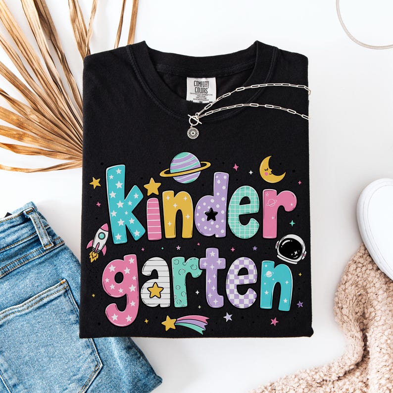 May include: A black t-shirt featuring the word kindergarten in large, multi-colored, patterned letters. The design includes space-themed graphics such as a planet with rings, a crescent moon, a rocket ship, an astronaut helmet, and stars.