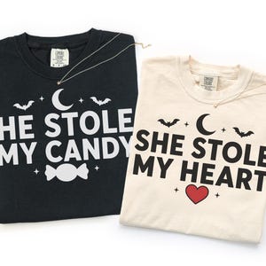 May include: Two crew neck t-shirts. The black shirt features white text reading HE STOLE MY CANDY with a crescent moon, bats, and a candy graphic. The cream shirt features black text reading SHE STOLE MY HEART with a crescent moon, bats, and a red heart.