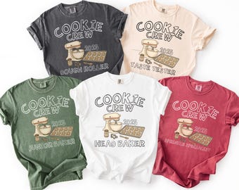 Peut inclure: Cinq t-shirts à manches courtes de couleurs variées : charbon, pêche, blanc, vert et rouge. Chaque t-shirt affiche "COOKIE CREW 2025" avec un dessin de mixeur et un titre lié à la pâtisserie : Dough Roller, Taste Tester, Junior Baker, Head Baker, et Sprinkle Specialist.