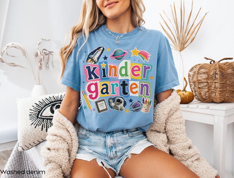 May include: A light blue t-shirt featuring the text "KINDERGARTEN" in colorful, patterned block letters. The design includes space-themed graphics like a rocket, planets, an astronaut, a pencil, a notebook, and a cup of crayons.