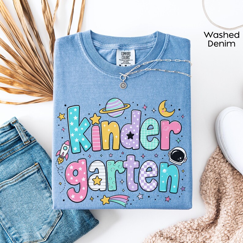 May include: A washed denim blue t-shirt featuring the text Kindergarten in large, colorful, patterned bubble letters. The design includes space-themed graphics such as a rocket, a planet with rings, a crescent moon, stars, and an astronaut helmet.
