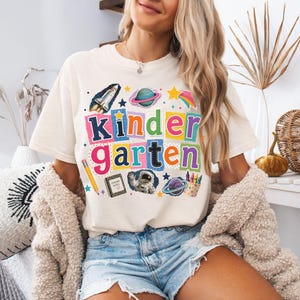 May include: An ivory t-shirt featuring a colorful graphic with the text "kindergarten" in block letters. The design includes a space shuttle, planets, a yellow pencil, a notebook labeled "teacher notes," an astronaut, and a cup of crayons.