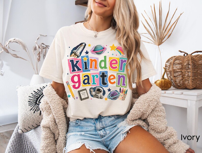May include: An ivory t-shirt featuring a colorful graphic with the text "kindergarten" in block letters. The design includes a space shuttle, planets, a yellow pencil, a notebook labeled "teacher notes," an astronaut, and a cup of crayons.