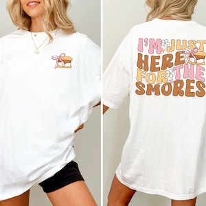 Op de afbeelding: Wit oversized T-shirt met een kleine s'mores-afbeelding aan de voorkant en de tekst "I'm just here for the smores" op de achterkant. De tekst is in roze, bruin en wit.