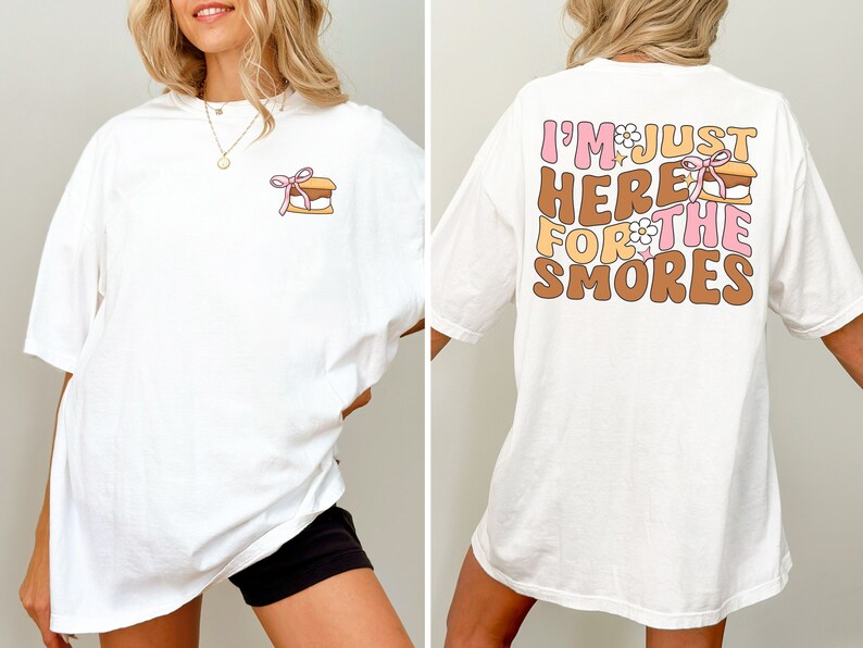 Op de afbeelding: Wit oversized T-shirt met een kleine s'mores-afbeelding aan de voorkant en de tekst "I'm just here for the smores" op de achterkant. De tekst is in roze, bruin en wit.