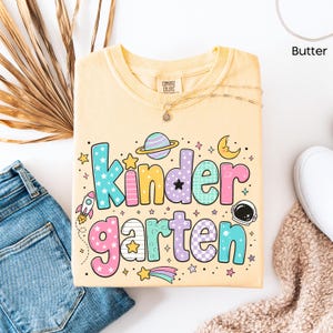 May include: A butter yellow t-shirt featuring the text Kindergarten in large, multi-colored, patterned bubble letters. The design includes space-themed graphics such as a planet with rings, a crescent moon, a rocket, and an astronaut helmet.