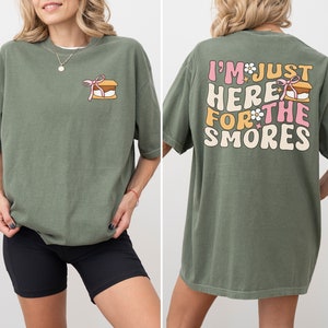 Op de afbeelding: Een groen T-shirt met een afbeelding van een s'more op de voorkant en de tekst "I'm just here for the smores" op de achterkant.