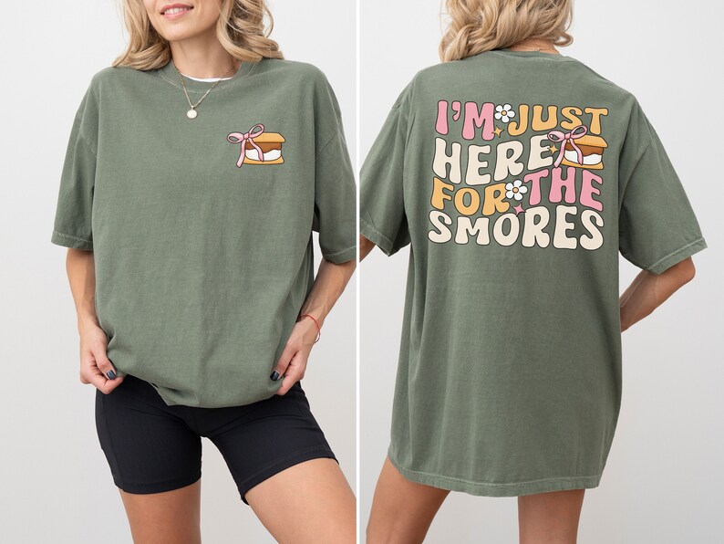 Op de afbeelding: Een groen T-shirt met een afbeelding van een s'more op de voorkant en de tekst "I'm just here for the smores" op de achterkant.