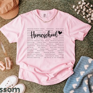 May include: Pink t-shirt with a black graphic that says "Homeschool" with a heart. The graphic lists many reasons why homeschooling is a good choice, such as "learning without limits", "creativity", "field trips", "faith", "personalized learning", "adventure", "real life lessons", "the world is our classroom", and more.