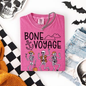 May include: A pink t-shirt with the words "BONE VOYAGE" and a graphic of dancing skeletons. The skeletons are wearing hats, sunglasses, and holding a cocktail, suitcase, and camera. A silver chain necklace is on the shirt.