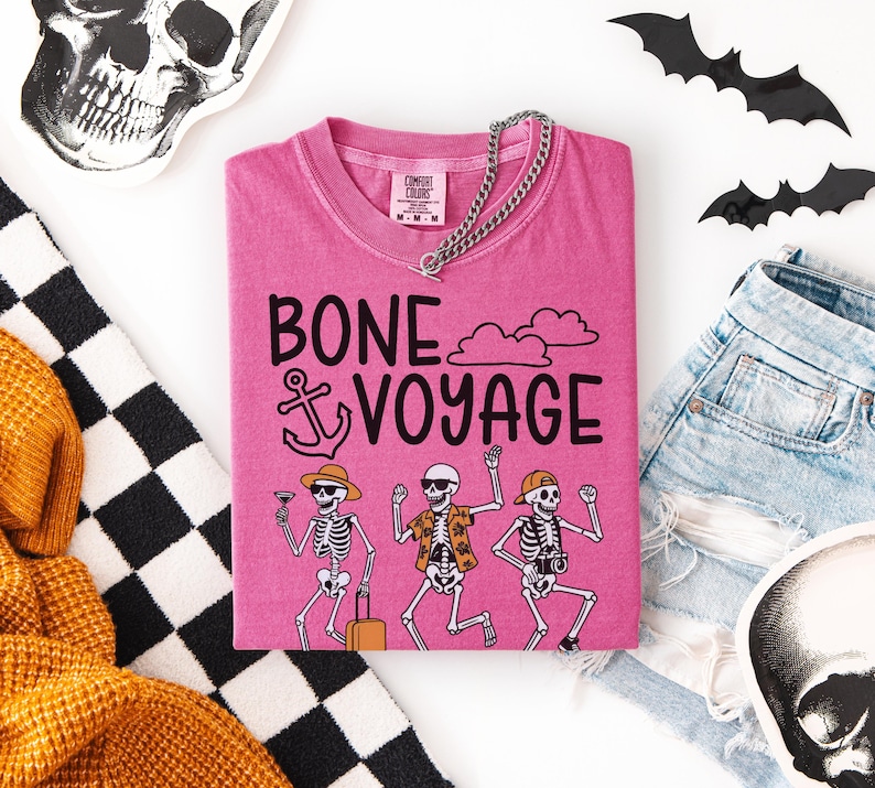 May include: A pink t-shirt with the words "BONE VOYAGE" and a graphic of dancing skeletons. The skeletons are wearing hats, sunglasses, and holding a cocktail, suitcase, and camera. A silver chain necklace is on the shirt.