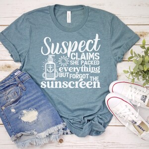 May include: A teal blue t-shirt with white text that reads "Suspect Claims She Packed Everything But Forgot The Sunscreen". The shirt features a graphic of a bottle of sunscreen with an anchor.