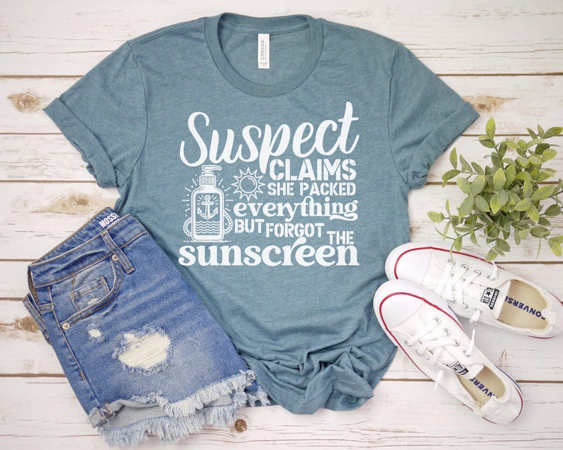 May include: A teal blue t-shirt with white text that reads "Suspect Claims She Packed Everything But Forgot The Sunscreen". The shirt features a graphic of a bottle of sunscreen with an anchor.