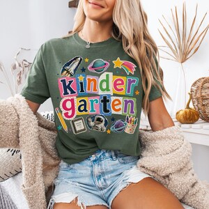 May include: An olive green t-shirt featuring a colorful graphic with the text "kindergarten" in multi-colored block letters. The design includes illustrations of a space shuttle, planets, a star, a pencil, a notebook, an astronaut, and crayons.