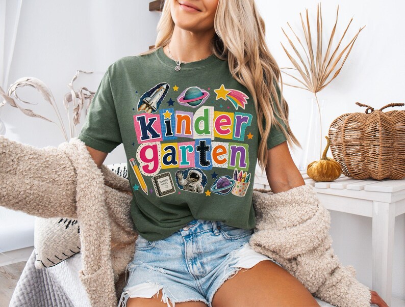 May include: An olive green t-shirt featuring a colorful graphic with the text "kindergarten" in multi-colored block letters. The design includes illustrations of a space shuttle, planets, a star, a pencil, a notebook, an astronaut, and crayons.