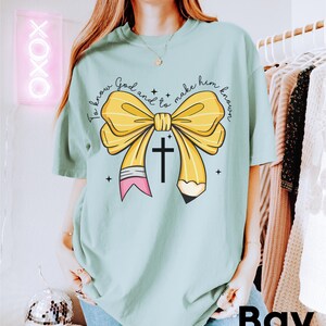 May include: A light blue t-shirt with a yellow pencil bow and a cross. The text on the shirt reads "To know God and to make him known."