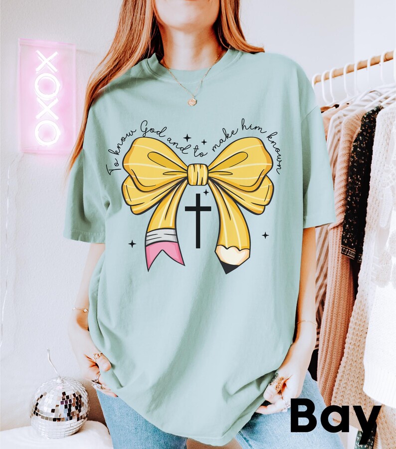 May include: A light blue t-shirt with a yellow pencil bow and a cross. The text on the shirt reads "To know God and to make him known."