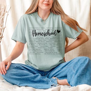 May include: A light blue t-shirt with a black graphic print that says "Homeschool" with a heart symbol. The graphic print includes a list of words related to homeschooling, such as "learning without limits", "family time", "discovery", "imagination", "real life", "confidence", "learning by doing", and "the world is our classroom".