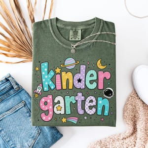May include: An olive green t-shirt featuring the text "kindergarten" in colorful, patterned bubble letters. The design includes space-themed graphics such as a planet with rings, a crescent moon, a rocket ship, an astronaut helmet, and stars.