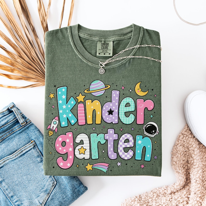 May include: An olive green t-shirt featuring the text "kindergarten" in colorful, patterned bubble letters. The design includes space-themed graphics such as a planet with rings, a crescent moon, a rocket ship, an astronaut helmet, and stars.