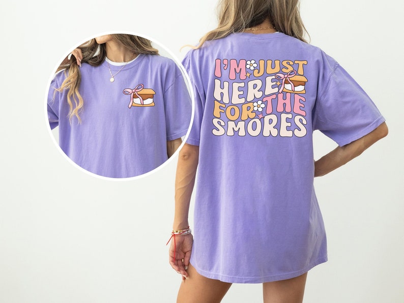 Op de afbeelding: Een paars T-shirt met de tekst "I'm just here for the s'mores" in roze en witte letters met een s'mores-afbeelding. Het shirt heeft een ontspannen pasvorm.