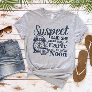 May include: A grey t-shirt with a navy blue graphic of an alarm clock and a coffee cup. The text on the shirt reads "Suspect said she would wake up early still asleep at noon".