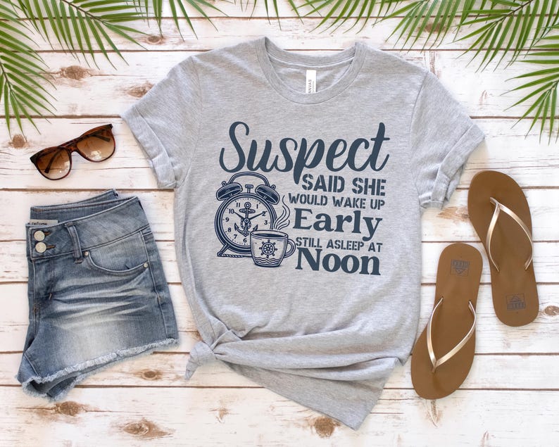 May include: A grey t-shirt with a navy blue graphic of an alarm clock and a coffee cup. The text on the shirt reads "Suspect said she would wake up early still asleep at noon".